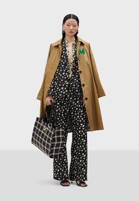 Hnedý dlhý kabát s zelenou nášivkou "M", blúzka a nohavice s čiernymi a krémovými bodkami, čierno-krémová kockovaná kabelka, čierne sandále na pásikoch.