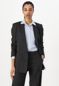 Black pinstripe suit jacket with double buttons worn over a light blue striped shirt. Features tailored fit and structured design.