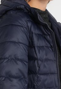 Tmavě modrá prošívaná bunda s kapucí, s prošívanými částmi, zapínáním na zip a hladkým lesklým povrchem.