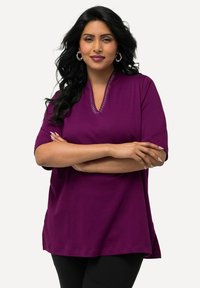 Túnica de algodón morada con mangas cortas, escote en V y detalles de costura decorativa en el cuello. Se lleva con pantalones negros.