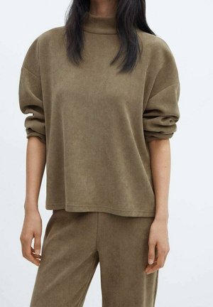 Femme portant un haut ample en velours côtelé marron à manches longues retroussées et un pantalon assorti sur un fond clair uni.