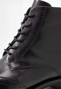 Botín de cuero negro con punta redonda, intrincados detalles de brogue y cordones planos. Textura suave con acabado pulido y suela resistente.