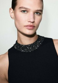 Top negro sin mangas con un cuello texturizado y brillante. La tela parece acanalada, y el escote presenta un detallado diseño de lentejuelas negras.