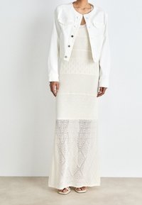 Valkoinen farkkutakki valkoisen, kuvioidun maksimekon päällä, jossa on pitsimäisiä tekstuureja ja monimutkaisia geometrisia kuvioita. Paksut, vaaleat sandaalit.