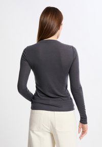 Donna di spalle che indossa una camicia a maniche lunghe aderente di colore grigio scuro e pantaloni bianco sporco con tasche posteriori, su uno sfondo chiaro.