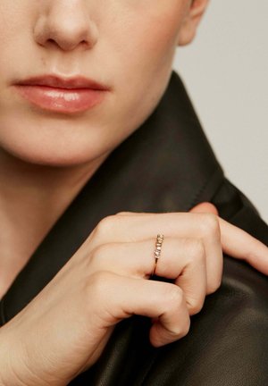 Anillo de oro con cinco piedras preciosas rectangulares, montado en una banda delgada. La mano descansa sobre una superficie de cuero negro, mostrando un tono de piel rosa claro.