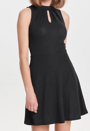 Femme portant une robe noire texturée sans manches avec un petit détail d'encolure en forme de fente (trou de serrure) et une jupe évasée.