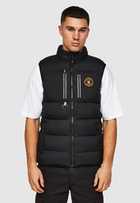 Zwarte puffervest met hoge kraag, voorrits, twee met ritssluiting afgesloten borstzakken en een rond logo; gedragen over een witte t-shirt en donkere broek.