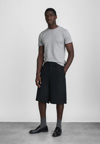 Muž v šedém tričku, černých kraťasech ke kolenům, šedých ponožkách a černých polobotkách, stojící na pozadí čistě bílé stěny.