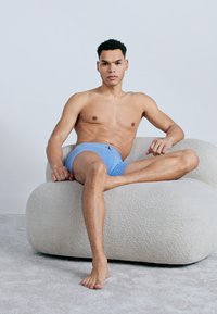 Άνδρας που φοράει ανοιχτό μπλε μποξεράκια κάθεται σε μια στρογγυλεμένη επενδεδυμένη καρέκλα με το ένα πόδι λυγισμένο και το άλλο τεντωμένο σε χαλιά δάπεδο.