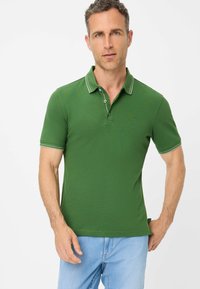 Polo verde con maniche corte, rifiniture bianche al colletto e alle maniche, e una chiusura a due bottoni. Indossato con jeans in denim azzurro chiaro.