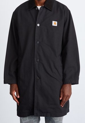 Homme portant un manteau Carhartt noir à boutons avec un col noir et des poches latérales, debout devant un fond uni.