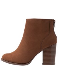 Brązowy zamszowy botek za kostkę z zaokrąglonym noskiem, bocznym zamkiem i masywnym, paskowanym obcasem. Gładka tekstura o jednolitym, solidnym kolorze.