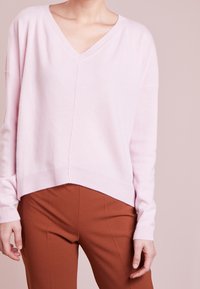 Jasnoróżowy sweter w serek z miękkiego dzianiny, z opuszczonymi ramionami i ściągaczem, zestawiony z dopasowanymi brązowymi spodniami. Casualowy, luźny krój.