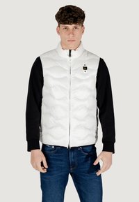 Hvid quiltet puffervest med høj krave, frontlynlås og et lille sort logo-mærke. Båret over en sort langærmet skjorte og blå jeans.