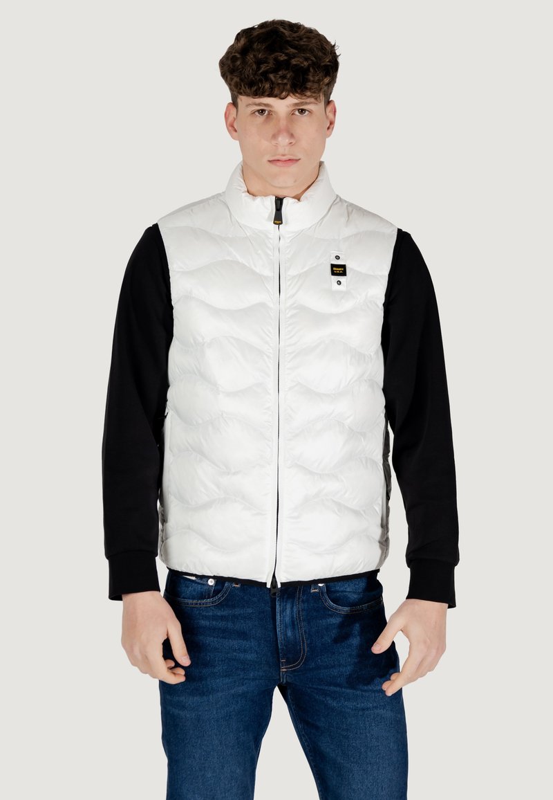 Hvid quiltet puffervest med høj krave, frontlynlås og et lille sort logo-mærke. Båret over en sort langærmet skjorte og blå jeans.