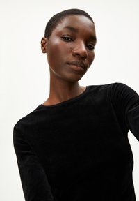 Schwarzes, samtiges Langarmshirt mit Rundhalsausschnitt, glatter Textur und taillierter Silhouette, präsentiert an einem Model mit kurzen Haaren.