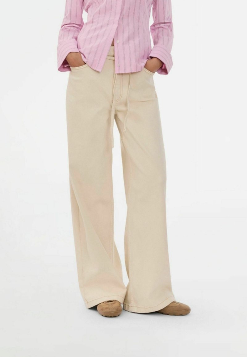 Osoba ubrana w beżowe spodnie o szerokich nogawkach i brązowe buty, z rękami w kieszeniach, w różowej koszuli na guziki z długim rękawem.