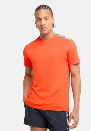 Orange kortærmet t-shirt i en tætsiddende design. Har kontrasterende sort tekst på skulderen. Blødt stof med en glat overflade.