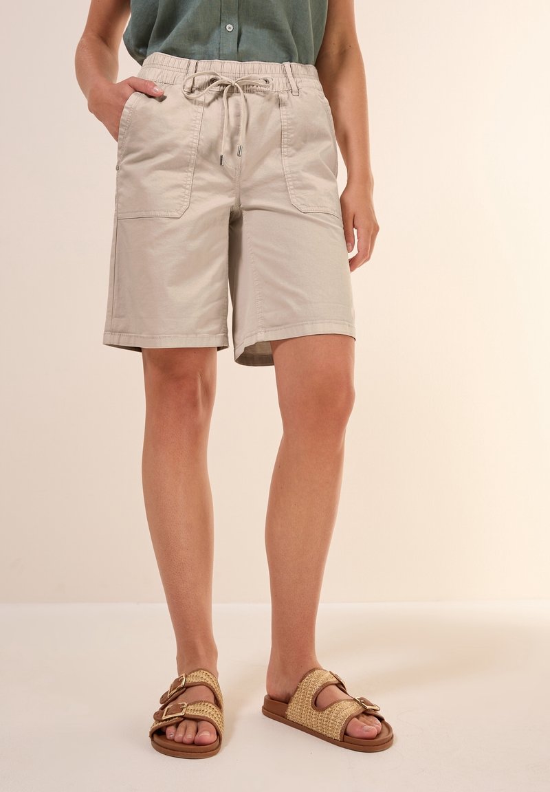 Personne portant un short beige à cordon, un haut vert sans manches et des sandales beiges avec des boucles, debout sur un fond clair.