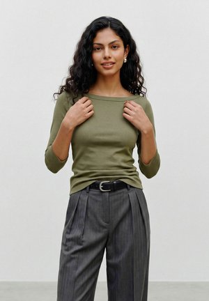 Jeune femme aux cheveux noirs bouclés porte un haut vert olive à manches longues, un pantalon plissé gris et des boucles d'oreilles en perles, debout devant un fond uni.
