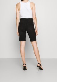 Shorts de mezclilla negros con dobladillo deshilachado, diseño ajustado y dos bolsillos traseros, combinados con una camiseta sin mangas blanca y sandalias de tacón negras.