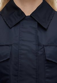 Κοντινή λήψη ενός ατόμου που φοράει μπλε σκούρο μπουφάν με μυτερό γιακά και δύο τσέπες στο στήθος με καπάκια.