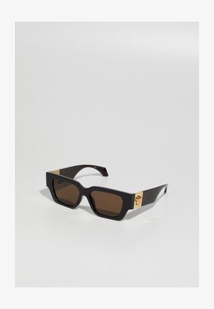Black sunglasses with a bold square frame, brown lenses, and gold accents on the arms featuring a logo detail. Smooth plastic texture.