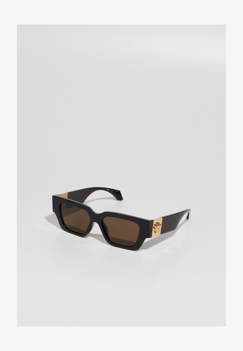 Mustad mustan värvi prillid, millel on julge ruudukujuline raam, pruuni läätsed ja kuldsed aktsendid käepidemetel, kus on logo detail. Sile plastikust tekstuur.