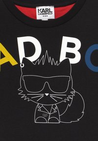 Camiseta negra para niños con letras coloridas "A D B C" y un contorno blanco de un gato con gafas de sol y chaqueta debajo de la etiqueta del cuello.