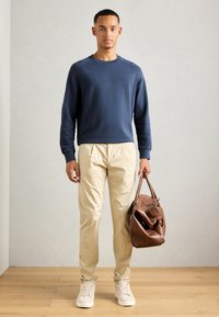 Young man standing wearing a navy sweatshirt, beige pants, white sneakers, and holding a brown leather bag in a neutral room.