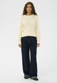 Pull en tricot crème avec un motif ajouré, associé à un jean large bleu foncé et des chaussures noires en maille à enfiler.
