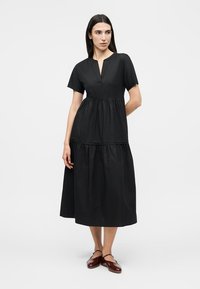 Γυναίκα με μακριά σκούρα μαλλιά που φοράει μαύρο midi φόρεμα με κοντά μανίκια και καφέ επίπεδα παπούτσια, στέκεται μπροστά σε ένα απλό φωτεινό φόντο.