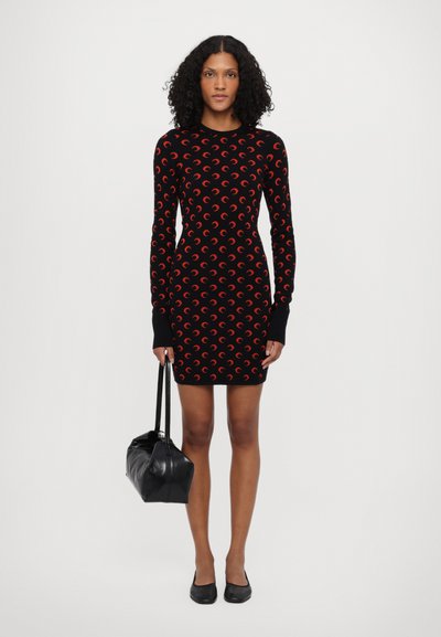 Vestido de punto negro con mangas largas, que presenta un patrón de luna creciente roja. La modelo lleva un bolso negro y calza unas bailarinas negras.