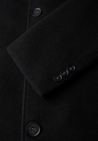 Schwarzer Wollmantel mit einem Reverskragen, vier schwarzen Knöpfen auf der vorderen Leiste und einem weichen, strukturierten Stoff.