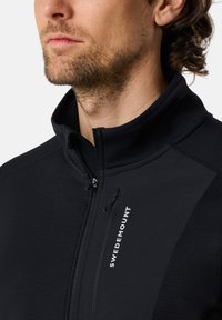 Black zip-up jacket with high collar. Features textured panels and a logo. Smooth material, fitted design, and front zip detail.