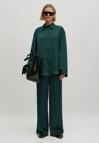 Grünes Satinhemd und weite Hose, kombiniert mit einer schwarzen lackierten Tasche. Das Model trägt übergroße schwarze Sonnenbrillen und spitze schwarze Schuhe.