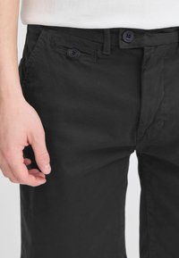 Gros plan d'une personne portant un pantalon noir avec une poche latérale boutonnée et un bouton de ceinture visible, main détendue le long du corps.