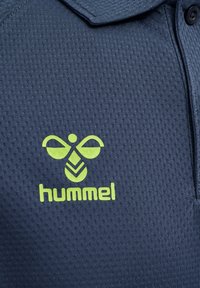 Polo shirt bleu marine en tissu texturé avec un logo vert d'une abeille stylisée et le nom de la marque "hummel" sur le côté gauche de la poitrine.