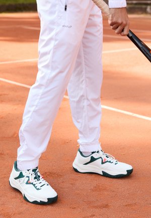 White athletic shoes with green accents, textured soles, and lace closure. Worn with white track pants, standing on a clay court.