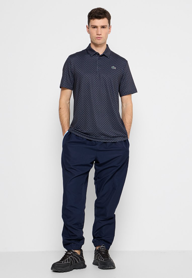 Polo shirt marine avec motif géométrique, manches courtes et col. Porté avec un pantalon de jogging marine assorti et des chaussures de sport noires.