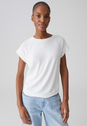 Jonge vrouw met een donkere huid en haar naar achteren gebonden, gekleed in een wit kortermig shirt en lichtblauwe spijkerbroek, staand tegen een neutrale achtergrond.