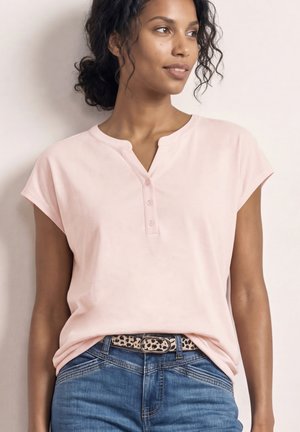 Kvinde med krøllet hår iført en lyserød bluse med korte ærmer og knapper, blå jeans og et leopardmønstret bælte, ser til siden.