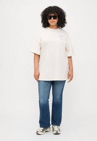 Kremfarget oversized t-skjorte med lilla grafikk, kombinert med blå slengjeans og svarte solbriller, med avslappet passform og glatt tekstur.