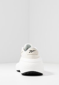 Beige träningsskor med vit texturerad sula, med en svart logotyp vid hälen och en ljusblå inredning. Rundad design.