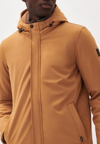 Chaqueta marrón, impermeable, con un alto cuello, capucha ajustable, bolsillos laterales y detalles de costura sutiles. Textura suave, diseño moderno.