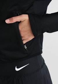Mano en el bolsillo con cremallera de una chaqueta negra texturizada, lleva pantalones Nike negros con el logo de swoosh blanco en la cintura.