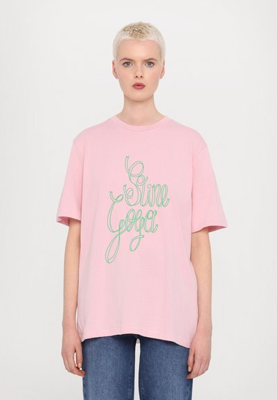 Rosa oversized t-skjorte med grønn kruset tekst "Sine Yoga". Laget av bomull med korte ermer og rund hals. Løs passform.
