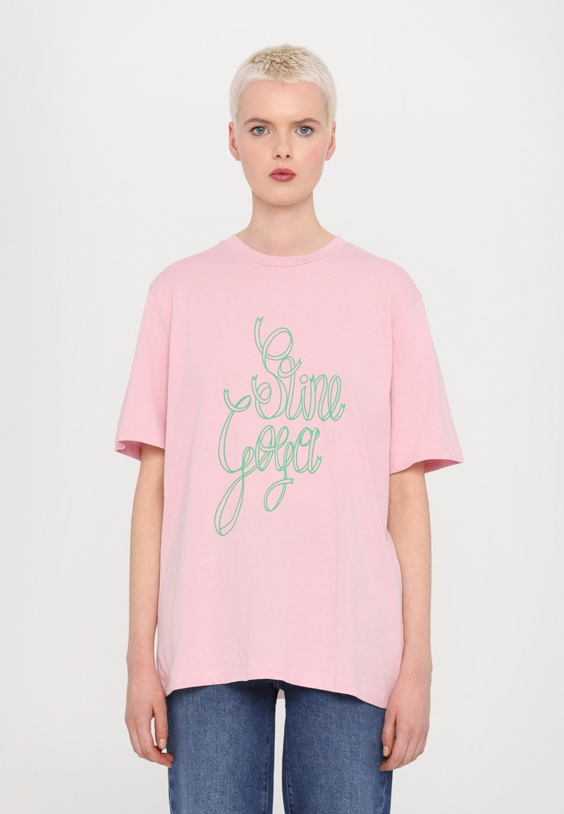 T-shirt oversized rosa com texto em verde cursivo "Sine Yoga". Feita de algodão, com mangas curtas e gola redonda. Corte casual.