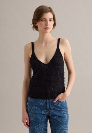 Donna con capelli corti che indossa un top nero senza maniche all'uncinetto e jeans blu con motivo paisley, in piedi davanti a uno sfondo semplice.